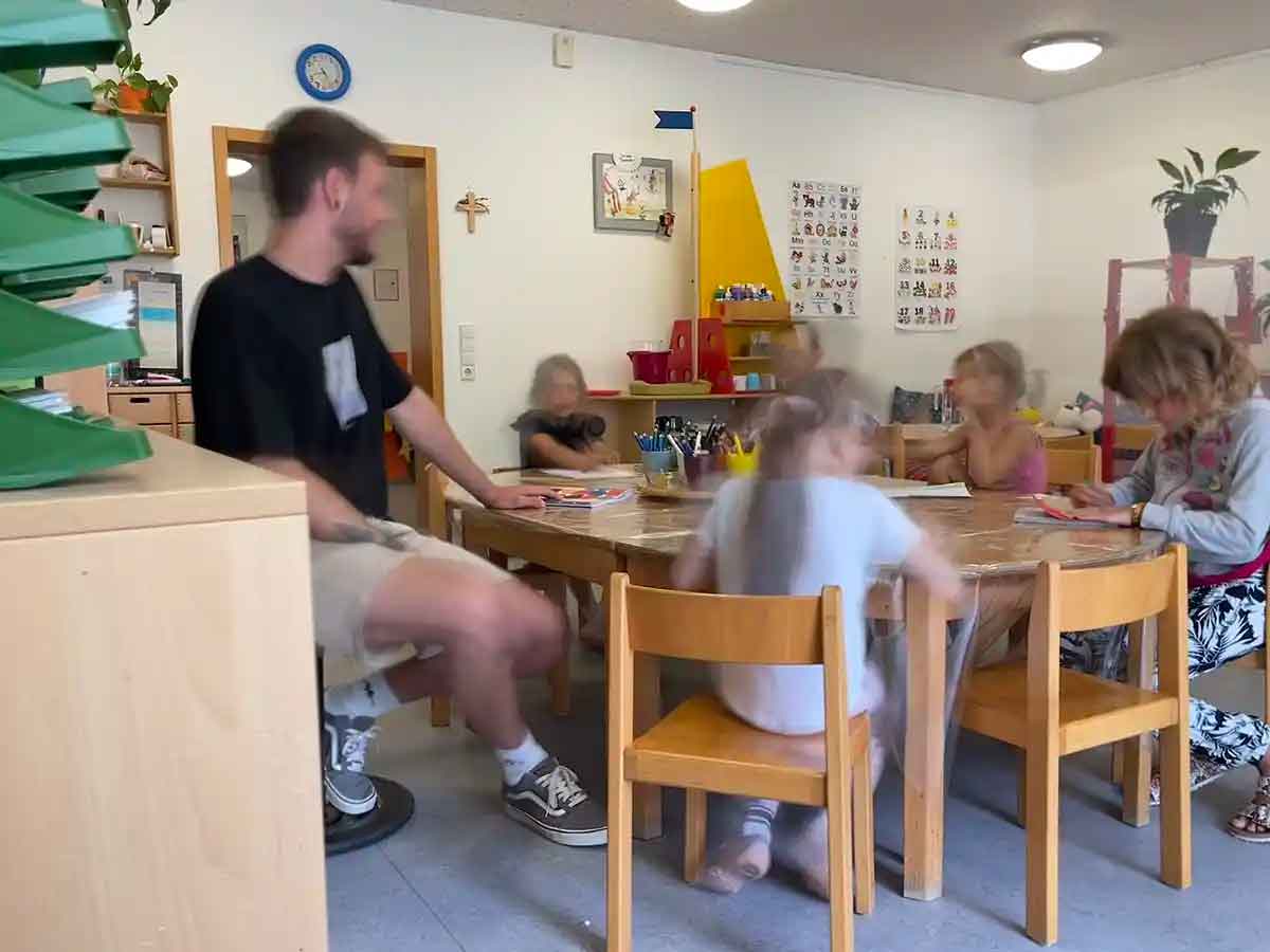 Im Kindergarten Bruder Klaus in Konstanz sitzt ein Erzieher an einem Tisch mit vielen Kindern. Auf dem Tisch liegt Mal- und Bastelmaterial.